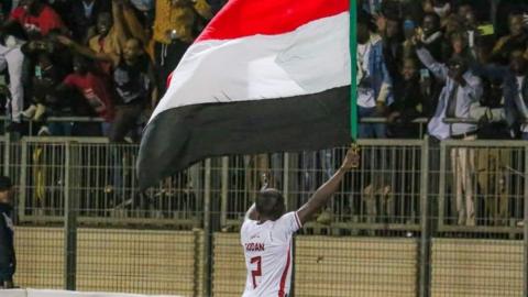 A Sudan player in a white shirt lifts a Sudanese flag ahead of supporters in the background