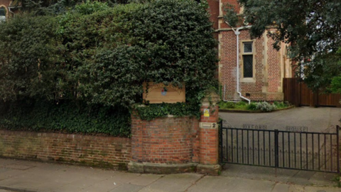 A brick wall and foliage behind it with a wooden sign with the words "Oxford House School" in front of a school building. To the right of the sign is a black metal gate with the words "Oxford House" on top.