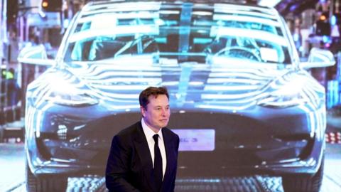 Elon Musk walking in front of a projected image of a Tesla car, wearing a black suit and tie.