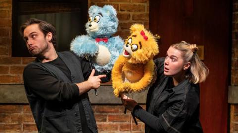 Two performers manipulate blue and yellow furry puppet characters during a scene from Avenue Q, with a brick wall set visible behind them.