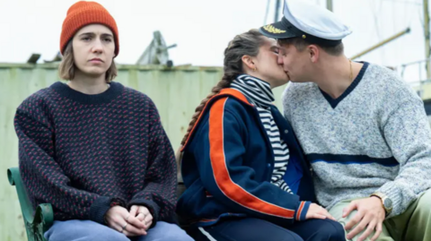 Three individuals sit closely on a bench in what appears to be a nautical setting—likely a dock or harbour area.

Left person: Wearing an orange beanie, a dark sweater with red and blue patterning, and light blue trousers. Their posture is relaxed, facing slightly toward the others.

Middle person: Dressed in a navy jacket with bold orange sleeve stripes over a striped shirt and dark trousers. They are engaged in a kiss with the person to their right.

Right person: Sporting a captain’s hat, a grey sweater with blue stripes, and light green trousers. They lean in toward the kiss, contributing to a moment of intimacy.

Behind them is a green wooden structure, possibly part of a boathouse or maritime equipment, with parts of a vessel in view—amplifying the scene’s coastal, ship-adjacent atmosphere.

There's a casual, candid charm here—like a still from a quirky indie film or a vintage postcard from an eccentric seaside story.