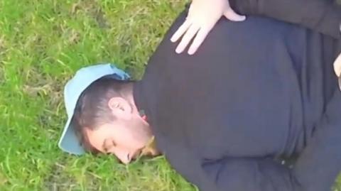 A man wearing a light blue cap is on the grass with his face turned to the side. He is being held down by a police officer (out of sight). Only hands can be seen.