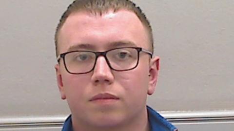A police mugshot of Cameron Miller. He is a young man with short light brown hair and black rimmed glasses.