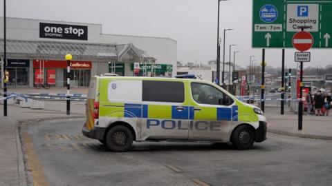 Police van on a street, in front of a cordon around part of Perry Barr. Police tape is wound around lamp-posts, with a shopping area in the background.