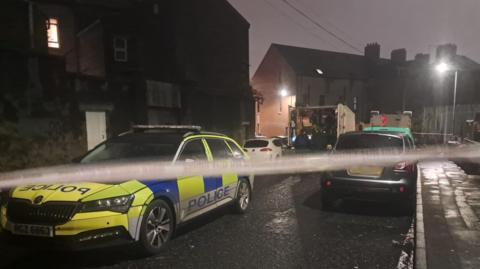 A street with police cordon, police car and other vehicles parked.