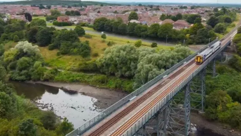 A train made up of two carriages cross a railway viaduct. Behind it you can see a large town. 