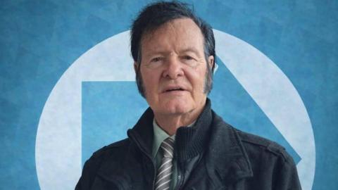 Victor Hagan has dark black hair and wears a green shirt and striped tie in this Reform Party profile