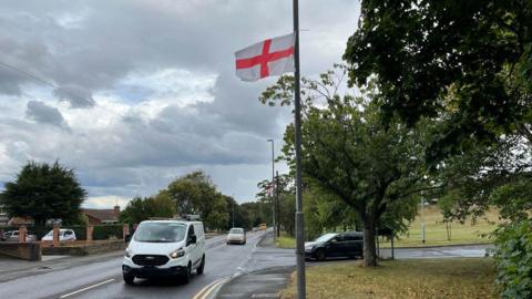 A St George's Cross flag which has been hung to a lamppost next to a junction in Gateshead. Another flag can be seen on a lamppost further down the road. A white van is driving past while a car is waiting to leave the junction.