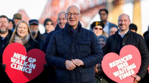 SNP leader John Swinney, a bald man wearing glasses and a blue padded coat, campaigning alongside a group of party colleagues holding heart-shaped pledge cards about the SNP's achievements in office - including "reduced NHS waiting times" and "new GP walk-in clinics"