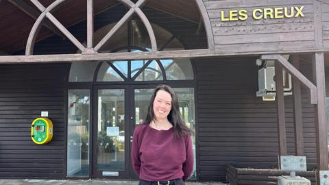 Jennifer Heald pictured outside Les Creux pavilion. She is wearing a plum coloured top and and is smiling. She has dark brown hair. In the background, the entrance to Les Creux is visible. The pavilion is mostly made of wood.