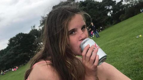 Holly Newton, who has long windswept brown hair, sits in a park drinking from a silver-coloured can of Diet Coke while looking at the camera.