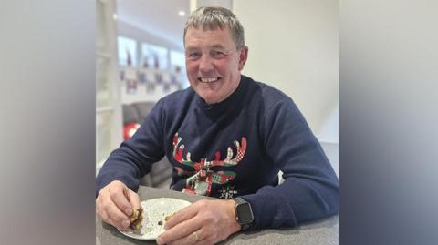 Richard Newson with short grey hair wearing a navy blue Christmas jumper featuring a tartan reindeer smiling and eating the remains of a mince pie