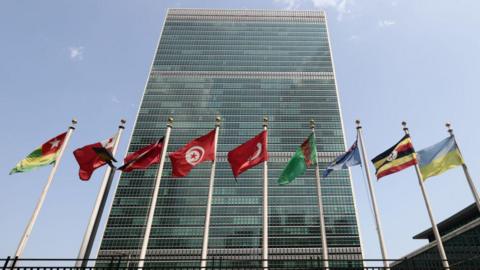 United Nations (UN) Headquarters in New York seen from outside. It is a tall tower block with lots of different countries' flags in front of it.