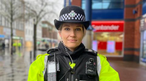 Police Sergeant Vicky Shears is stood on Slough high street. She is wearing a police uniform with a florescent vest under a black vest and a police hat bearing a police badge and a chequered black and white design. She has blonde hair. 