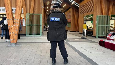 A police officer wearing black trousers and a black jacket which says POLICE on the back is standing in Great Yarmouth Market Place. The building is made of timber and features green metalwork gates to close the market off at night.
