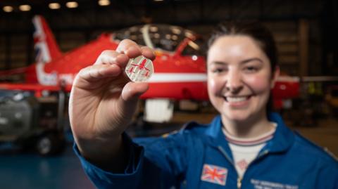 A young woman with brown hair tied back wearing blue overalls with a Union Flag badge smiling and holding up a 50p coin. It has three planes on it – the Red Arrows in red. Behind her is a Red Arrows plane.