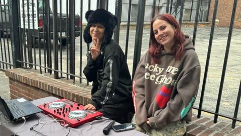 Two women in sweatshirts stand in front of a table with DJ equipment on a sidewalk