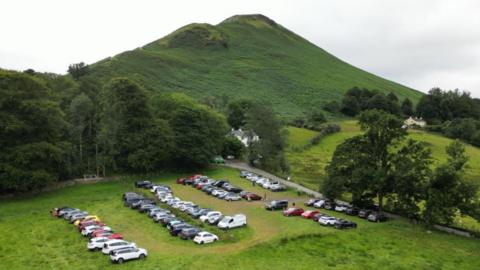 An aerial view of a field with dozens of cars parked in it, with Cat Bells fell visible beyond.