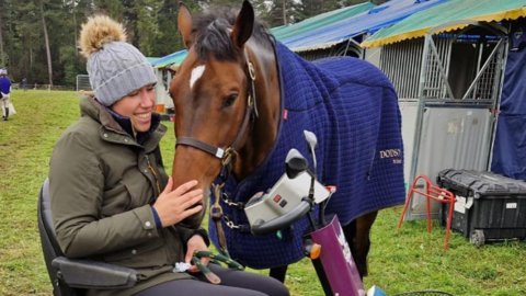 Caroline March in a green waxed jacket and grey bobble hat, who is sitting in a mobility scooter while stroking the nose of chestnut coloured horse. The horse is wearing a blue coat 