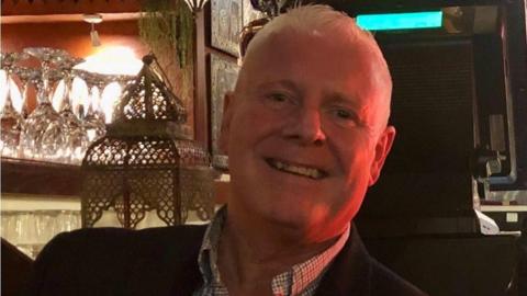 Peter Bartle smiling and wearing a checked shirt and black jacket, sat in what appears to be a restaurant as there are wine glasses behind him