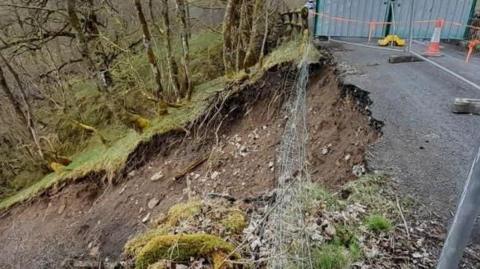 A close up of the landslip. A section of the carriageway has crumbled away leaving a steep slop of brown earth running to the left of the screen. A metal fence has been erected around the area to prevent people from coming too close to the slope.