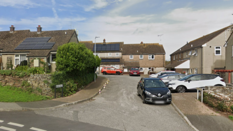 Google image of School Hill in Padstow. It is a residential street. Cars are parked in driveways outside of homes. One car is parked on the road next to the pavement. Two of the houses have solar panels on their roofs. A number of cones are place on the road, stopping motorists from parking.