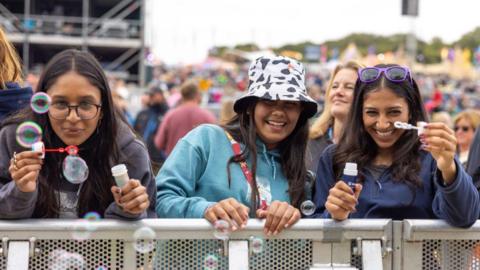 Three young women standing behind a barrier, with two on either side blowing bubbles from small tubs.