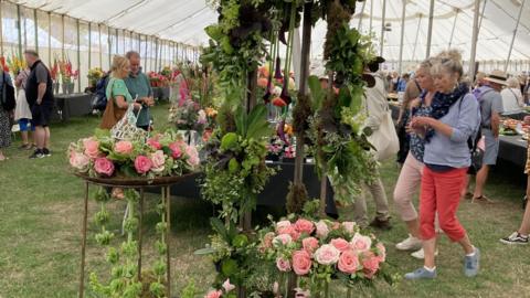 A large marquee with intricate flower displays inside. Two women are walking past one display which features roses of different shades of pink