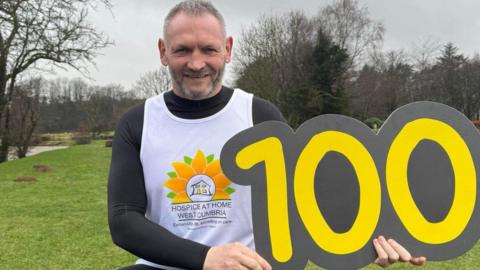 Gary McKee holding a large sign with the number 100 in yellow letters against a dark grey background. He has short grey hair and a close-cropped beard, is wearing running gear, and smiling. He is standing on a grassy bank, with trees and a glimpse of a river just to the left.