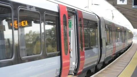 A close-up of one side of a white and grey train with one set of red doors open. It is parked next to a railway station platform.