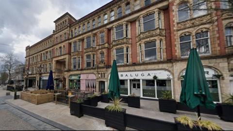 Street view of the Miller Arcade in Preston, a four-storey red brick building with a striking Victorian design with intricate details, large bay windows with stone surrounds and a mix of shops, restaurants and bars on the ground floor with seating outside. The top floor has much smaller windows surrounded by stone