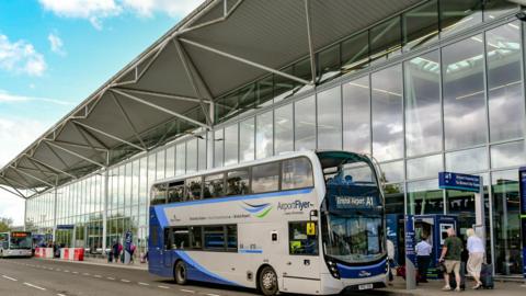People queueing for bus outside Bristol Airport, which has big glass windows and other buses in the distance.