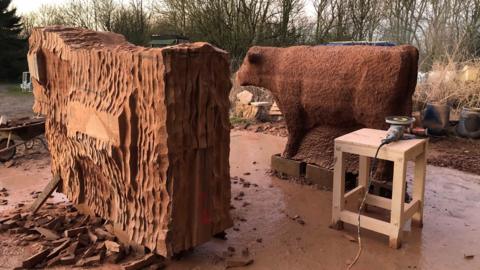 Two brownish-red sculptures placed outside. One is a bull with a shaggy coat and the other is a cow. You can see a circular electric tool that is plugged in and placed on a table next to the sculptures.