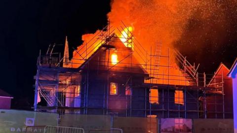 An under-construction house on fire,  with wood scaffolding in front of the house. Orange and pink flames can be seen inside the property.