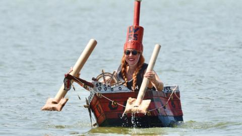 A woman is paddling in a cardboard boat on the water, wearing a cardboard hat.