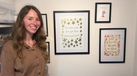 Kerri Cunningham is smiling at the camera in her office. She is standing in front of a cream wall where five of her sketches are hanging in black photo frames. One reads: 'All flowers bloom in their own time' while another reads: 'The world is so much brighter because you exist'.