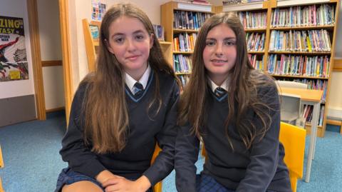 Two pupils at John Cabot Academy, sitting on yellow chairs in the library. They are both wearing a school uniform consisting of a blue and black checked skirt, navy jumper and dark tie. Both have brown hair and are sitting close together.