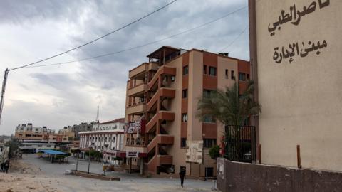 Brown buildings are seen under a cloudy sky, with a small figure walking past and debris on the ground