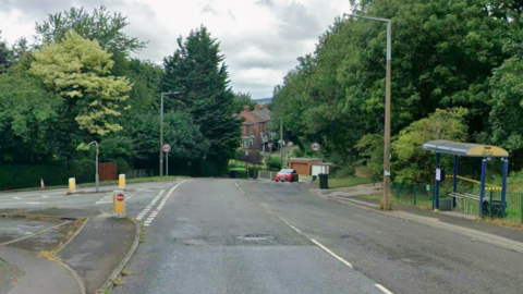 A Google Street View image of the junction of Redbrook Road and Beever Lane. To the right of the road is a bus stop. At the bottom of the hill a few houses are visible through the trees.
