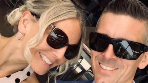 Emma and Lee Lovell smiling into the camera. Both are wearing black sunglasses. Emma has blonde hair which is in an up do with some hair framing her face. Lee has short dark hair. 