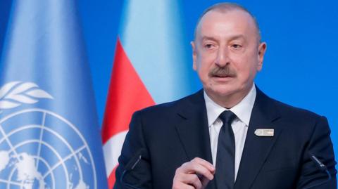 Azerbaijan's President Ilham Aliyev mid-speech at COP. He wears a dark blue suit and stands in front of a United Nations flag.