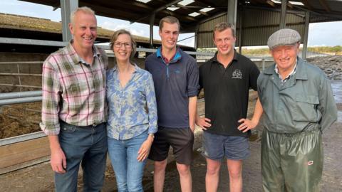 Andrew Jones with wife Jayne, sons Harvey and Oscar and father Roger are standing in a line in a barn. They are all smiling at the camera.