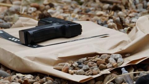 A black gun laid on a brown paper bag. It is being inspected by police. 
