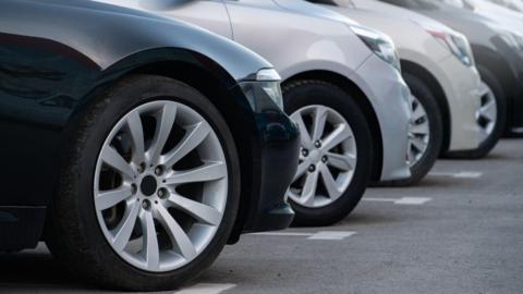 A closeup of the fronts of several cars parked pointing to the right of the image. The cars are a mixture of black, silver and white.