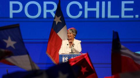Jeanette Jara stands at a podium with a blue backdrop, flanked by a Chilean flag.