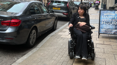 A woman in a wheelchair looking at a car parked in front of a dropped kerb.