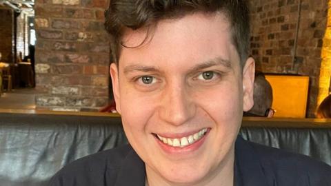 Head shot of Joel Shooter sitting on a lather seat in front of a brick wall smiling. He has wavy brown hair