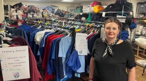 A woman stands at the front of a room packed with school uniform. Some are folded onto shelves at the back of the room and other are hung on rails.