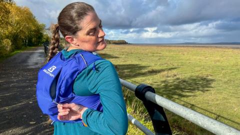 Gayle Redmon on a seaside trail twisting to look behind her while reaching with her right arm to open a zipper on a blue vest with a large pocket on the back