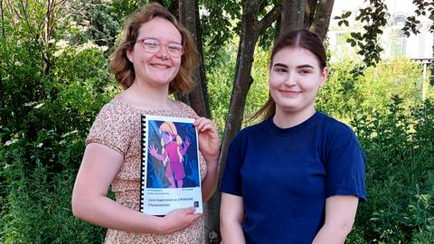 Rebecca and Fay, stood in front of a tree, Rebecca is holding a folder which contains a petition asking to make it illegal to discriminate against those in care